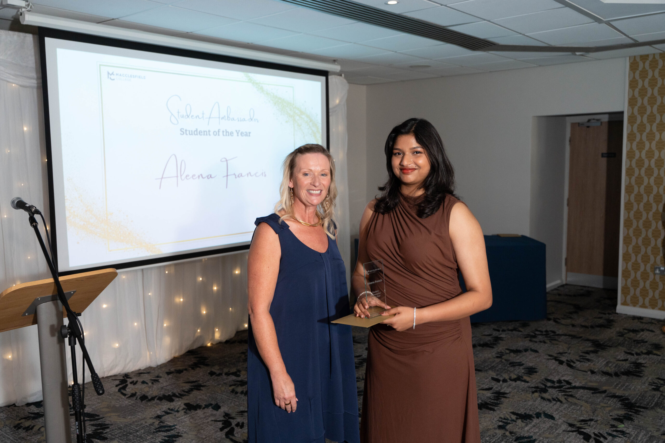 A female staff member and female student are smiling for a photo during a student awards event. The student is holding a glass award