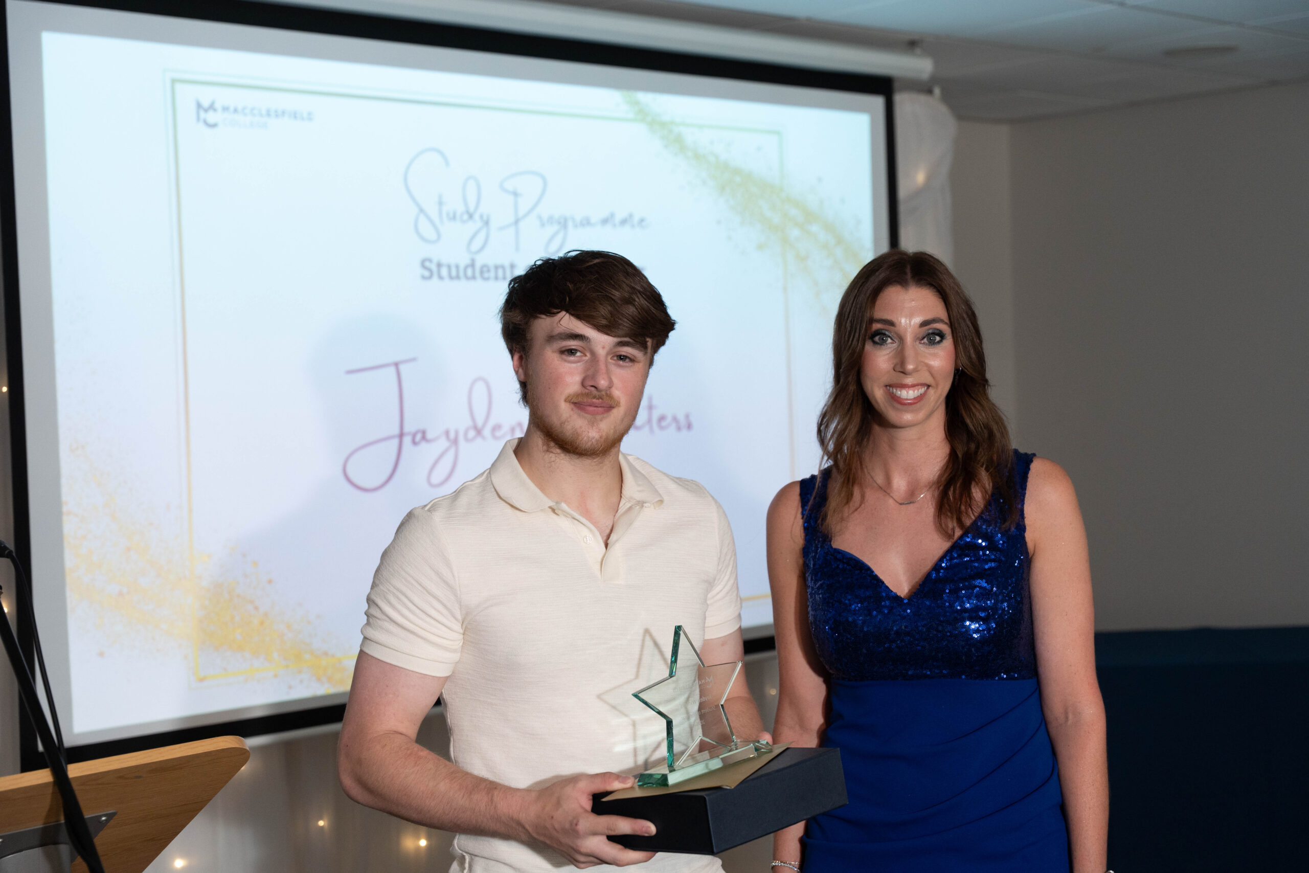 A female staff member and a male student are standing together posing for a photo. The student is holding a glass award