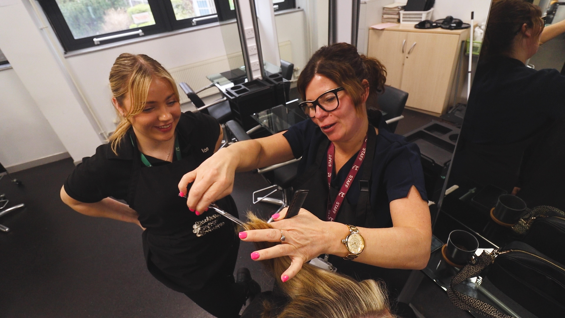 a hairdressing apprentice watches as a teacher demonstrates how to cut hair