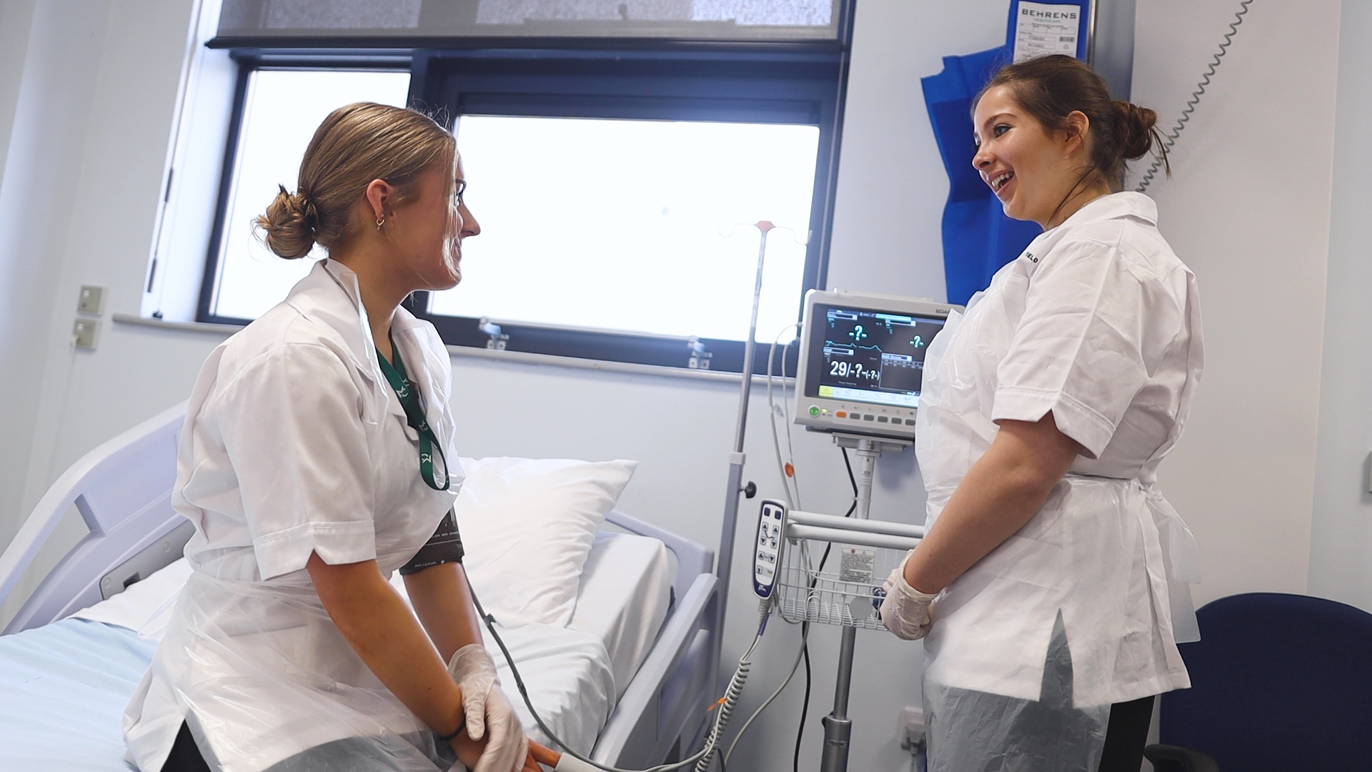 two health students, one sits on a bed while the other stands by a machine, are talking and smiling
