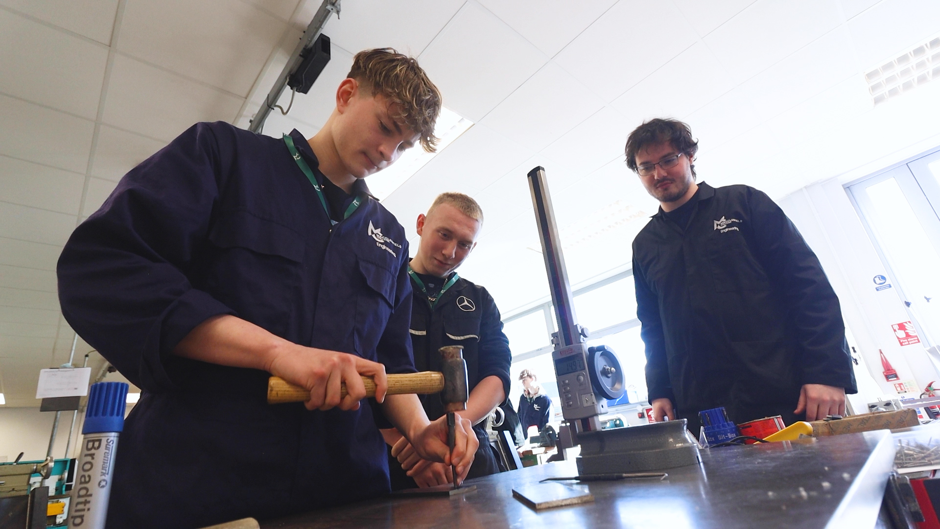 An engineering teacher and a student watches as another student is using a hammer
