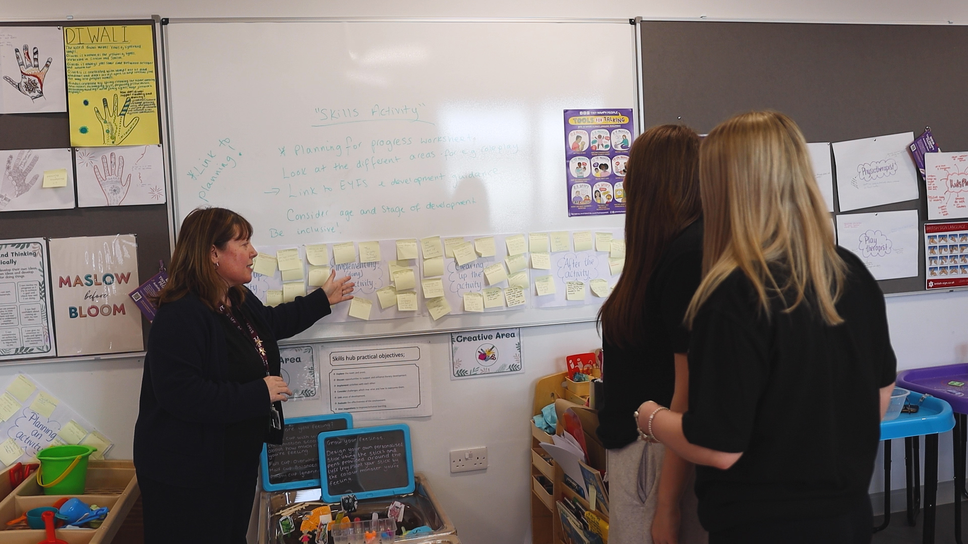 A childcare teacher and two students are looking at a board with sticky notes on it