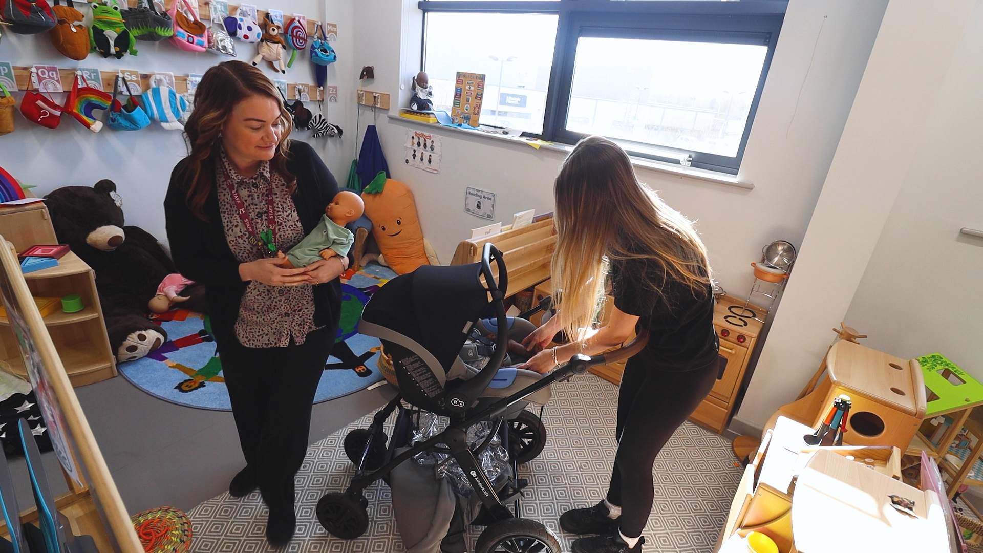 a childcare teacher holds a plastic baby whilst a childcare student is looking down at a pram