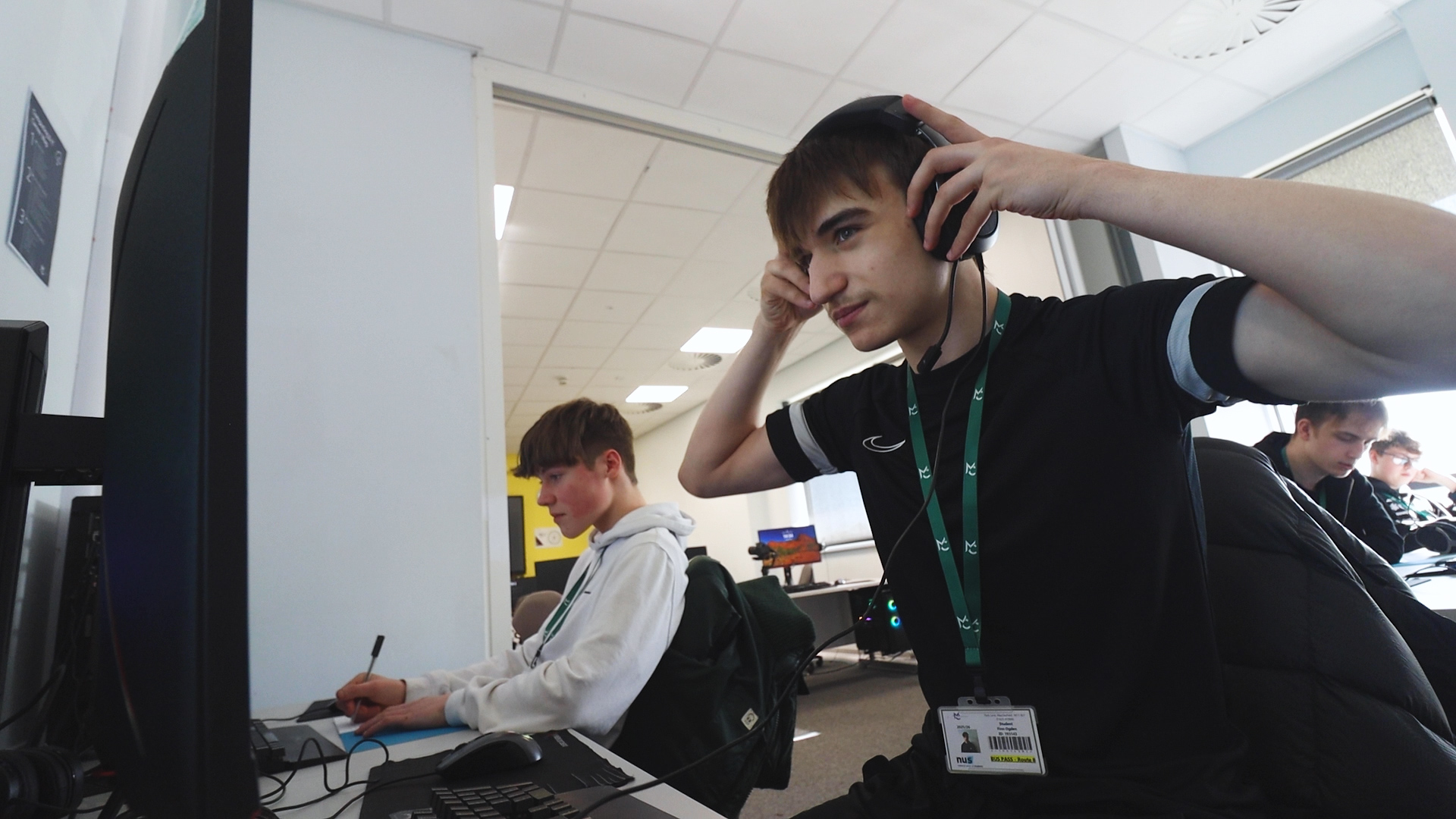 Two students are sitting working at computers. One student is putting on headphones