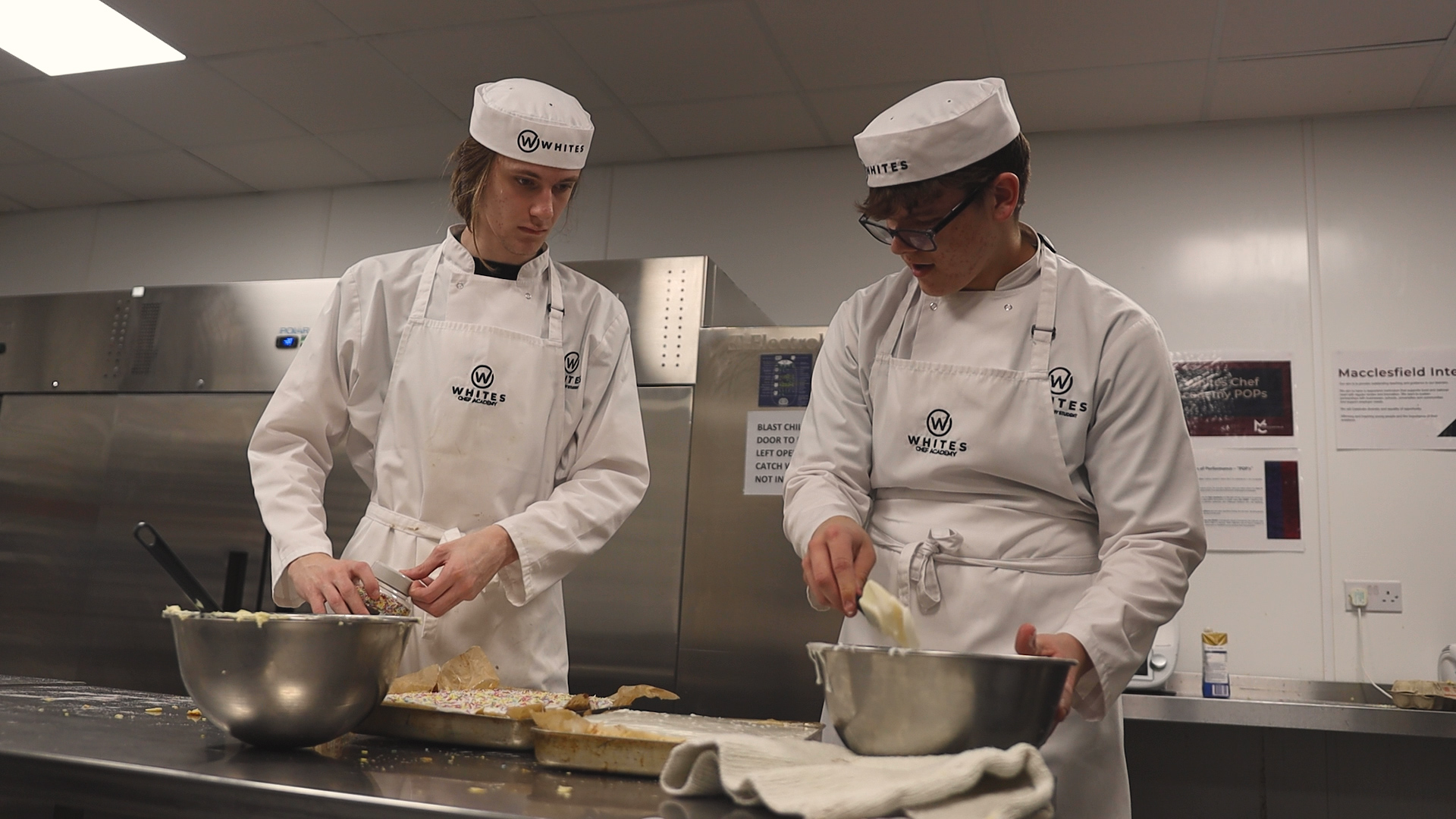 two students wearing chef uniforms are working in a kitchen at macclesfield college