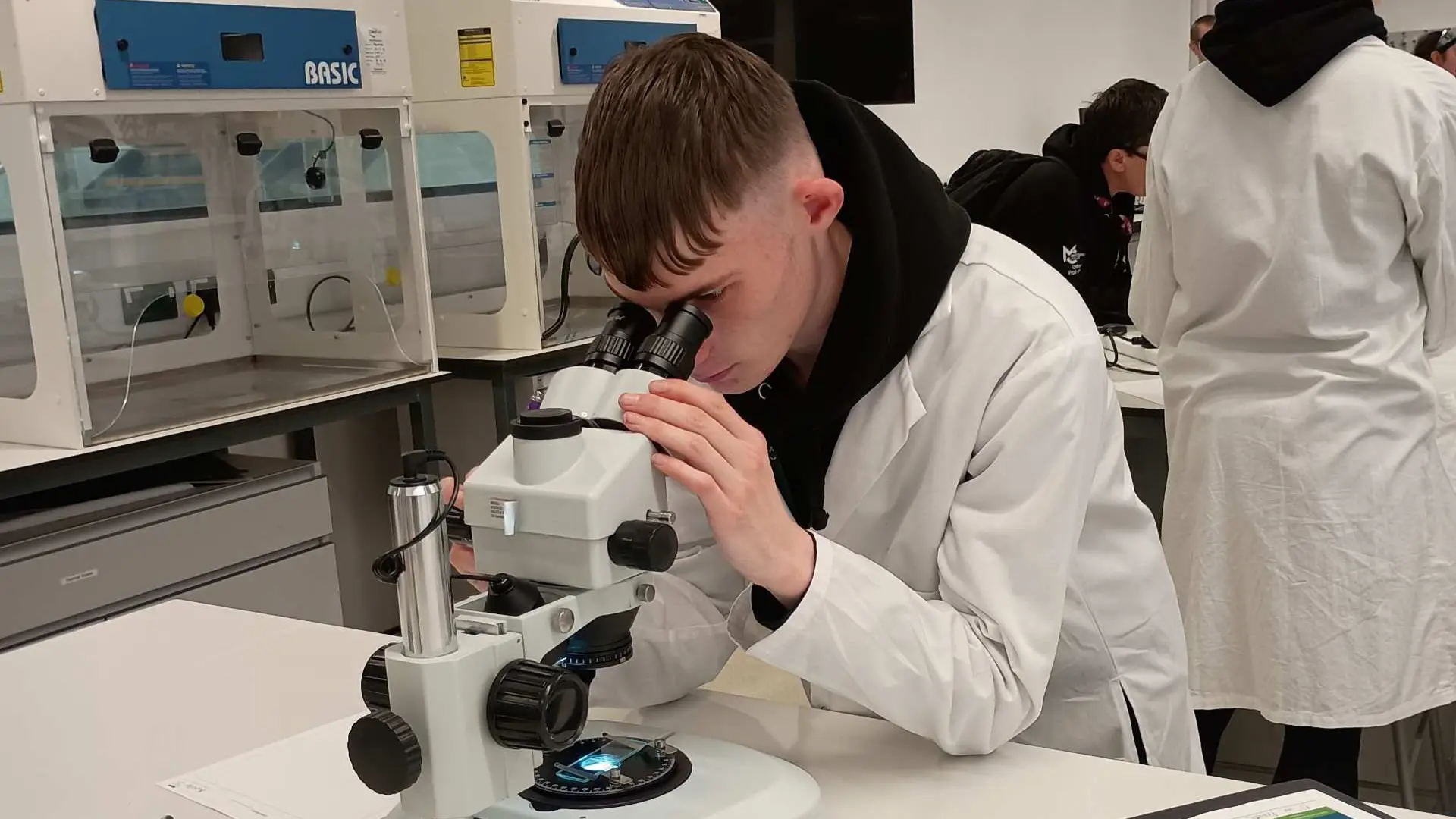 A public service student is wearing a lab coat and looking into a microscope