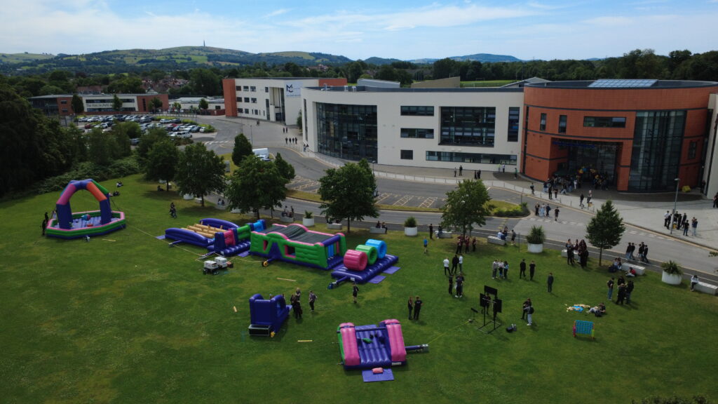 On a field in front of Macclesfield College there are a range of bouncy inflatable activities and a group of students standing near them.