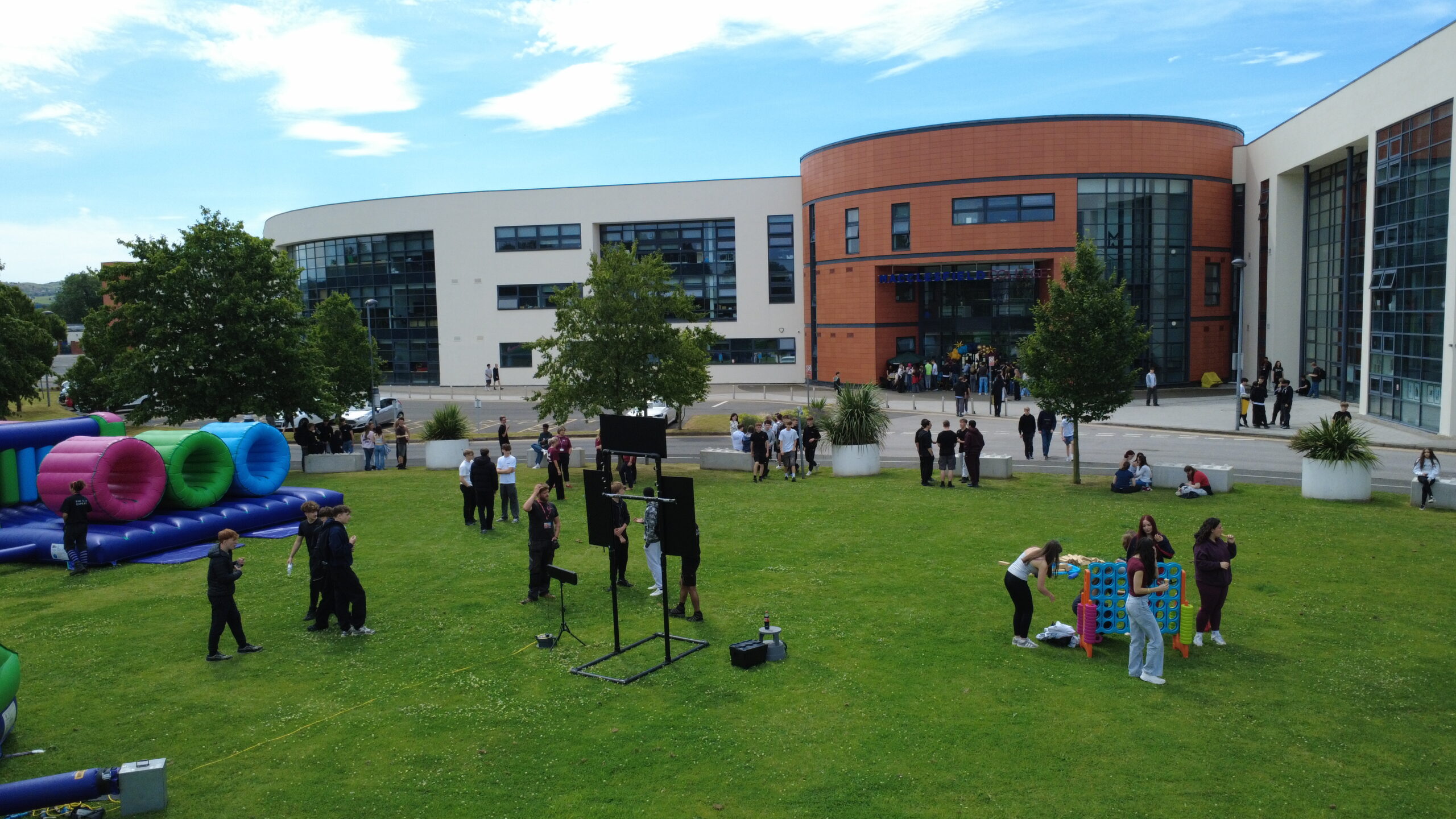 On a field in front of Macclesfield College there are a range of bouncy inflatable activities and a group of students standing near them.