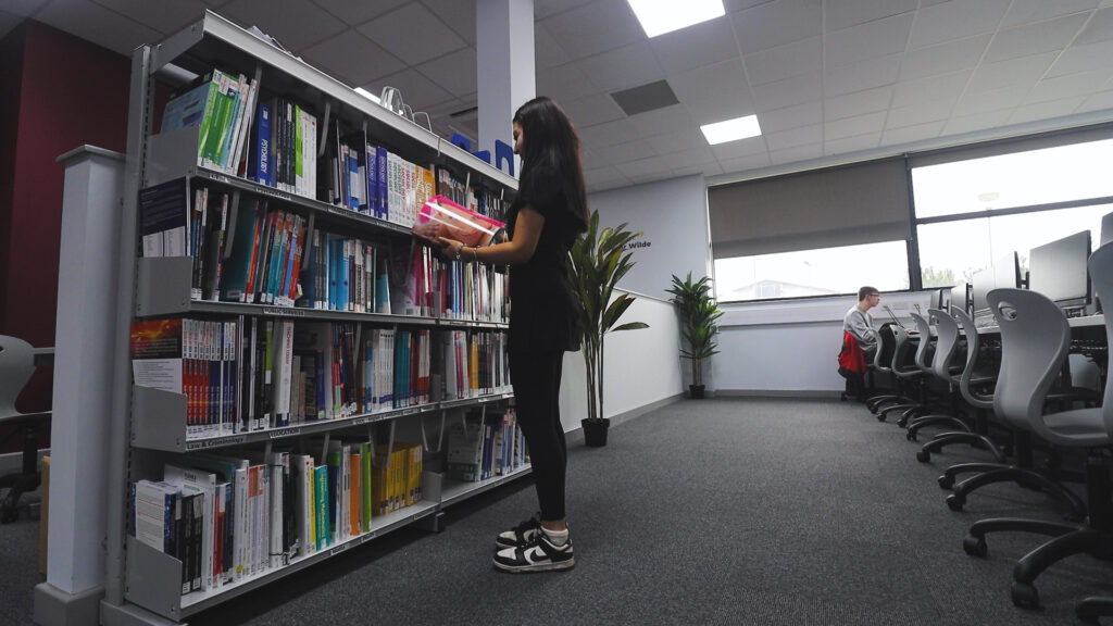 A female student is picking a book off a bookcase in the Student Hub