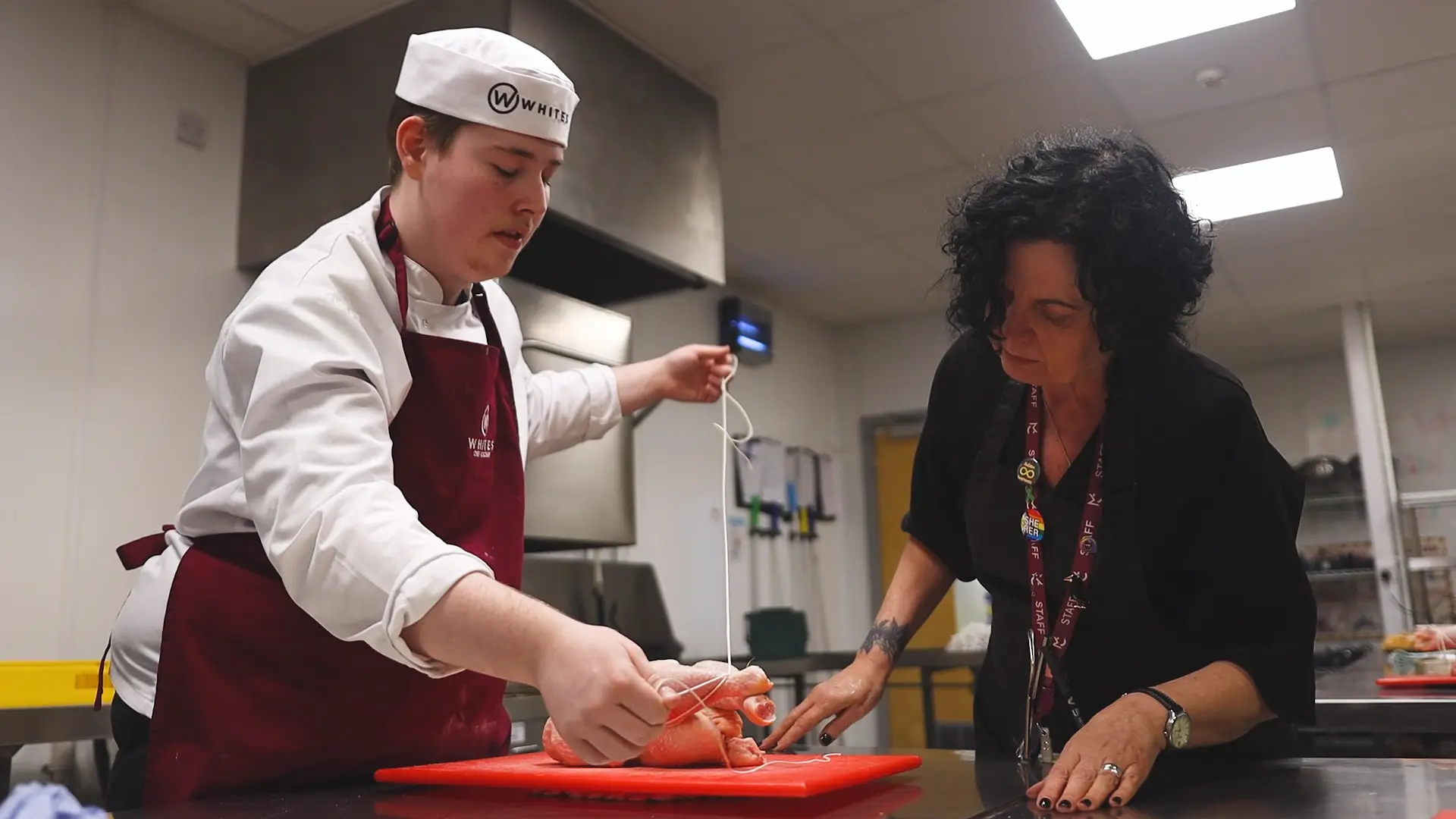 A whites chef academy student and teacher is putting string around chicken meat on a chopping board