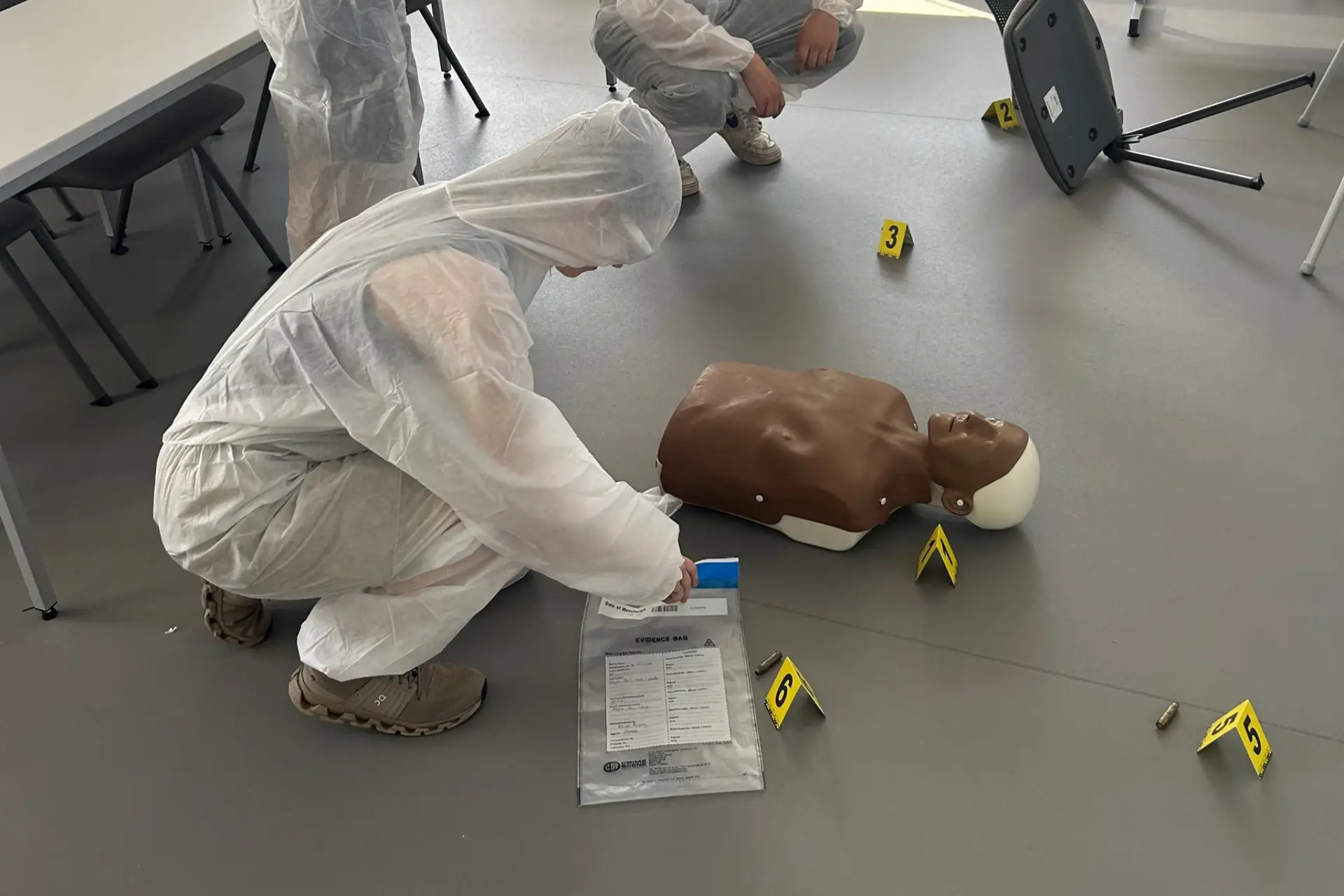 Three A level students are dressed in investigation scrubs while they examine a crime scene