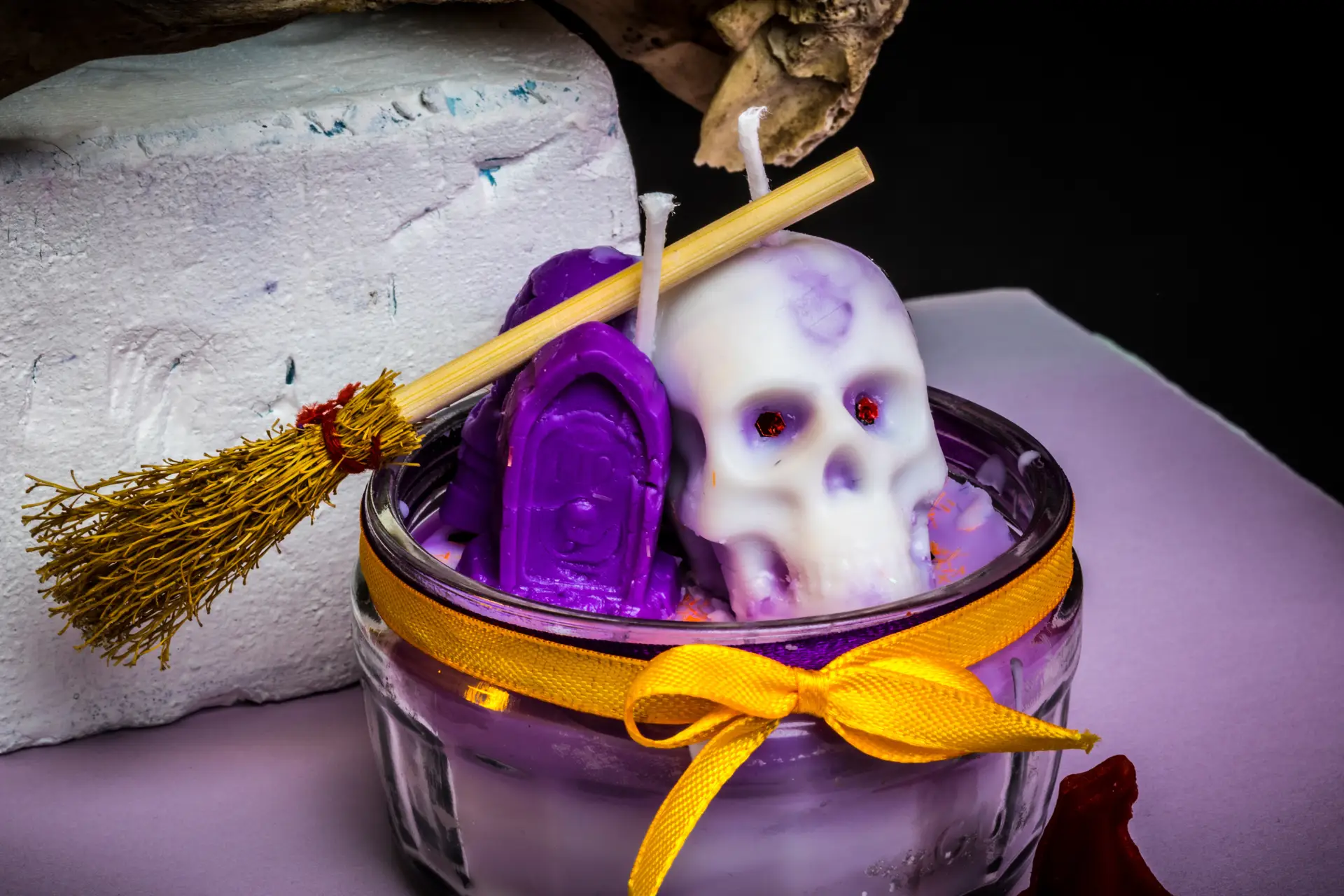 Glass jar candle decorated with a yellow ribbon, featuring a white skull shaped candle, purple gravestone candles, and a miniature broomstick.