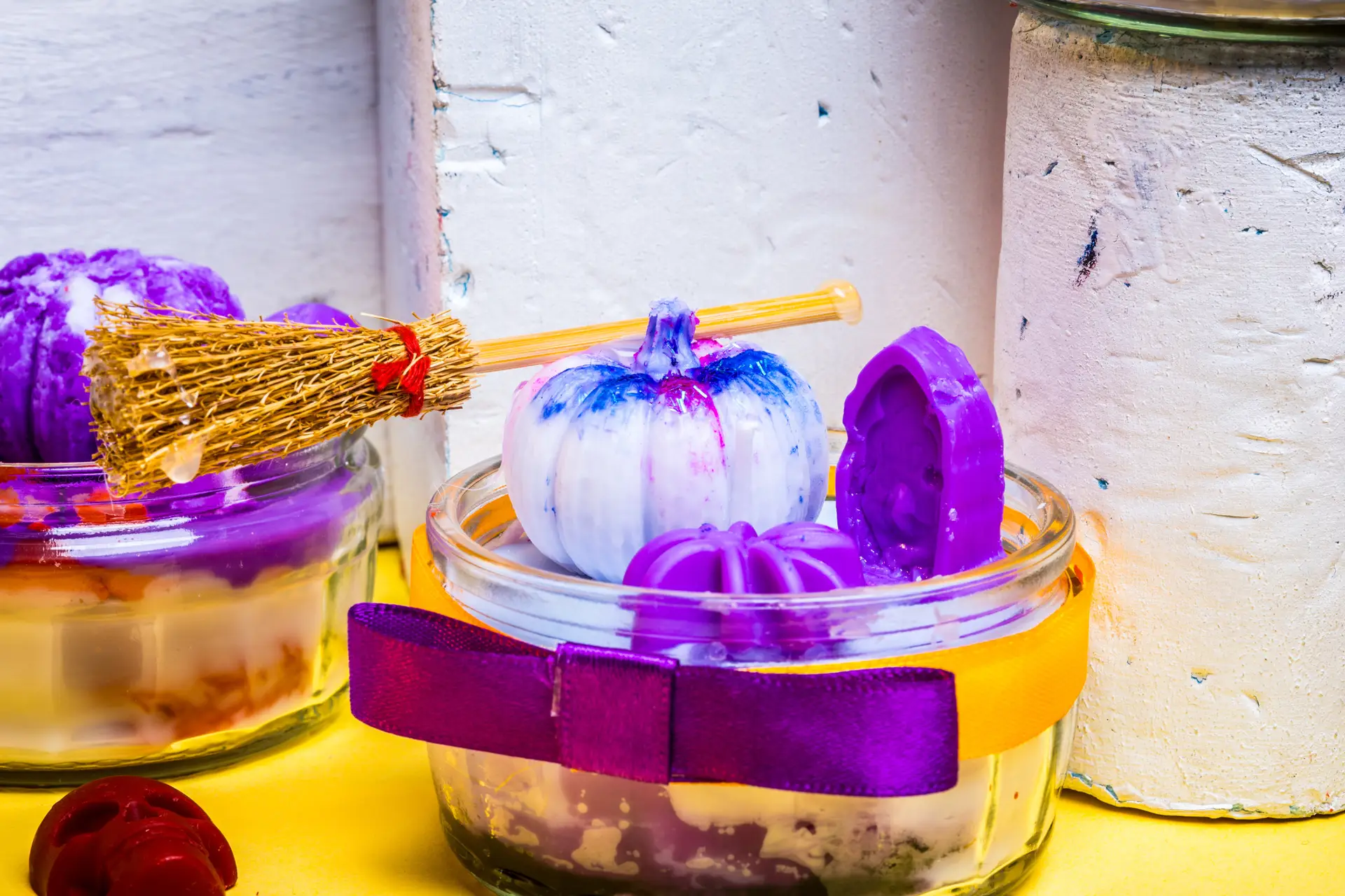 Glass jar candle decorated with a yellow and purple ribbon, featuring purple wax gravestone, purple wax spider, a mini pumpkin and a broomstick.