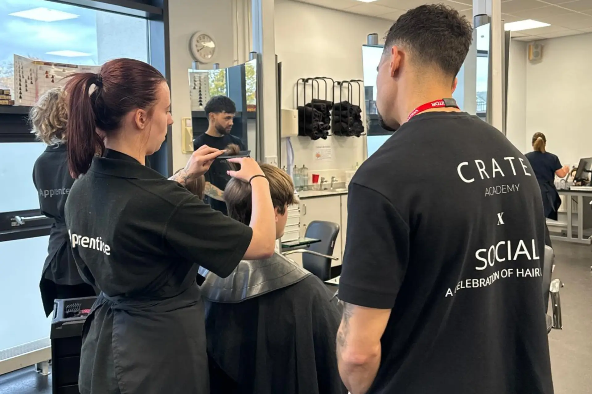 A hairdressing apprentice is practising barbering techniques while a male hairdresser watches.