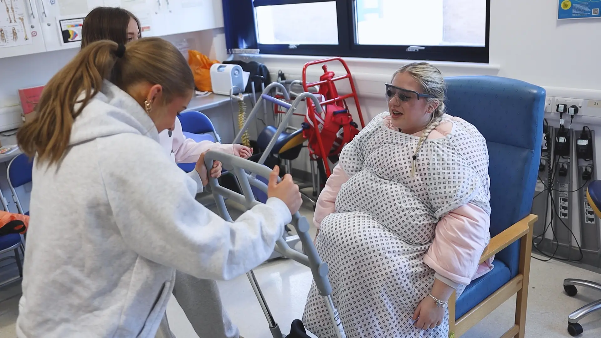 A female health student sits in a chair with a walking frame attached to it. She is wearing a fat suit. Next to her, two other health students stand.