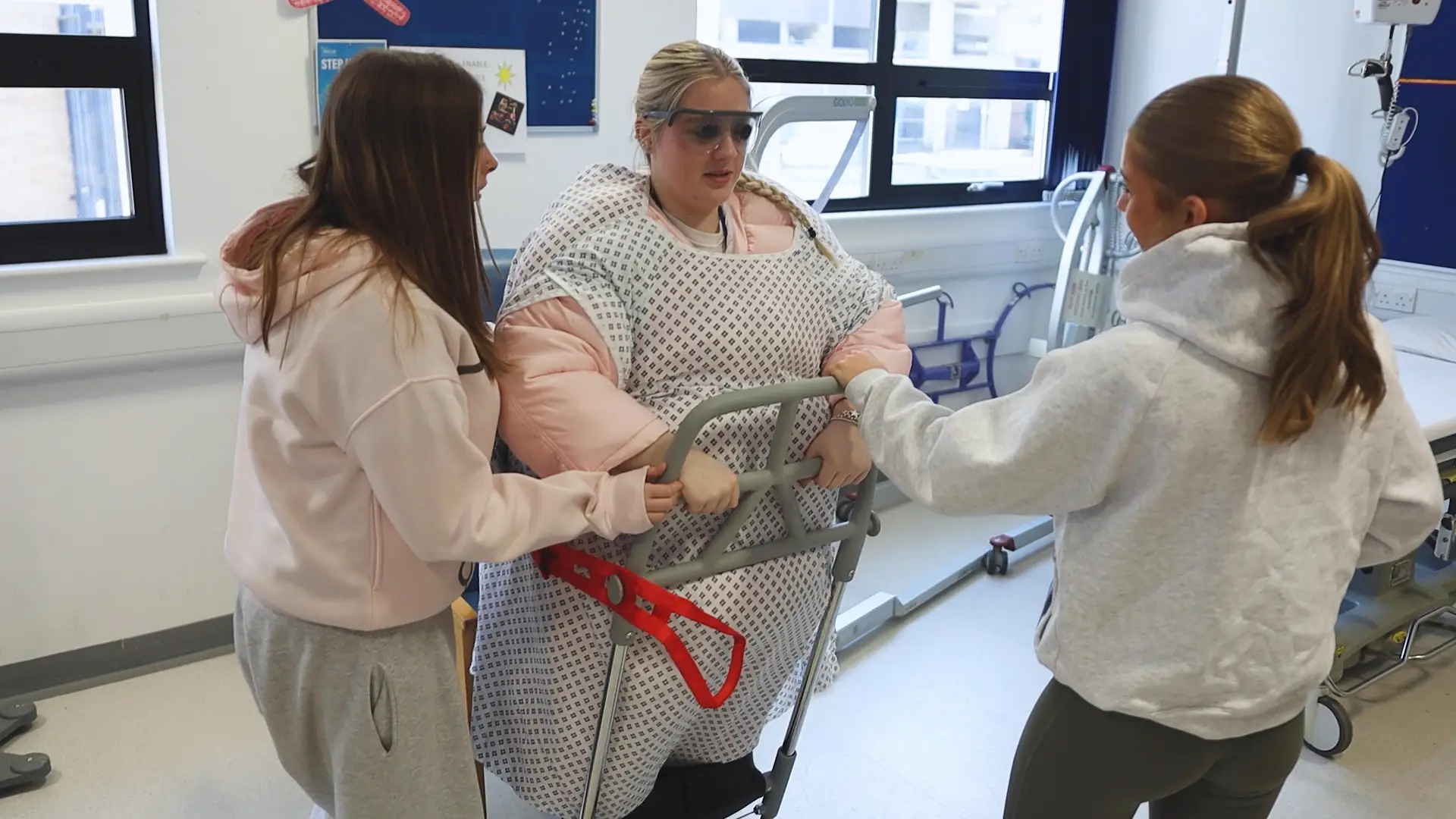 Two health students are helping another health who is dressed in a fat suit with a walking frame.