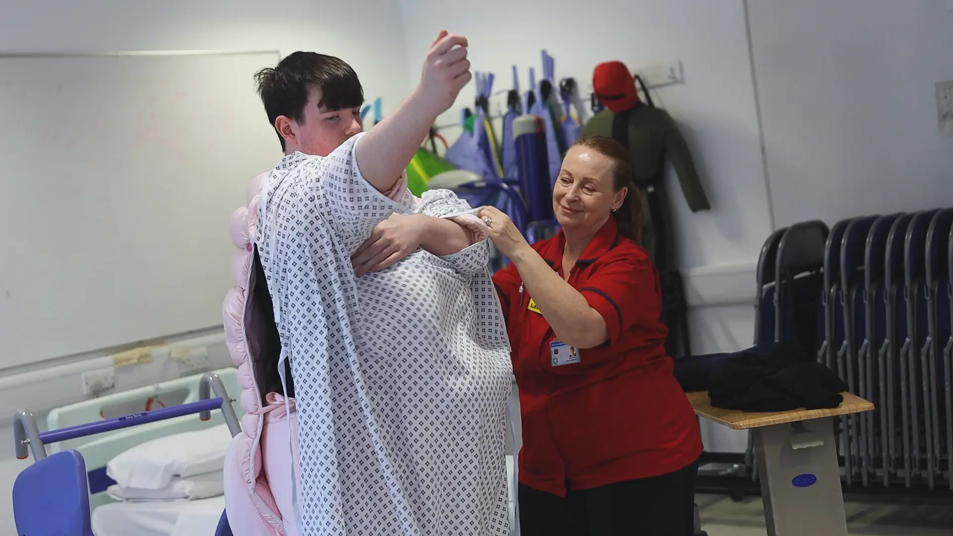 A hospital worker is helping a health student get into a fat suit.