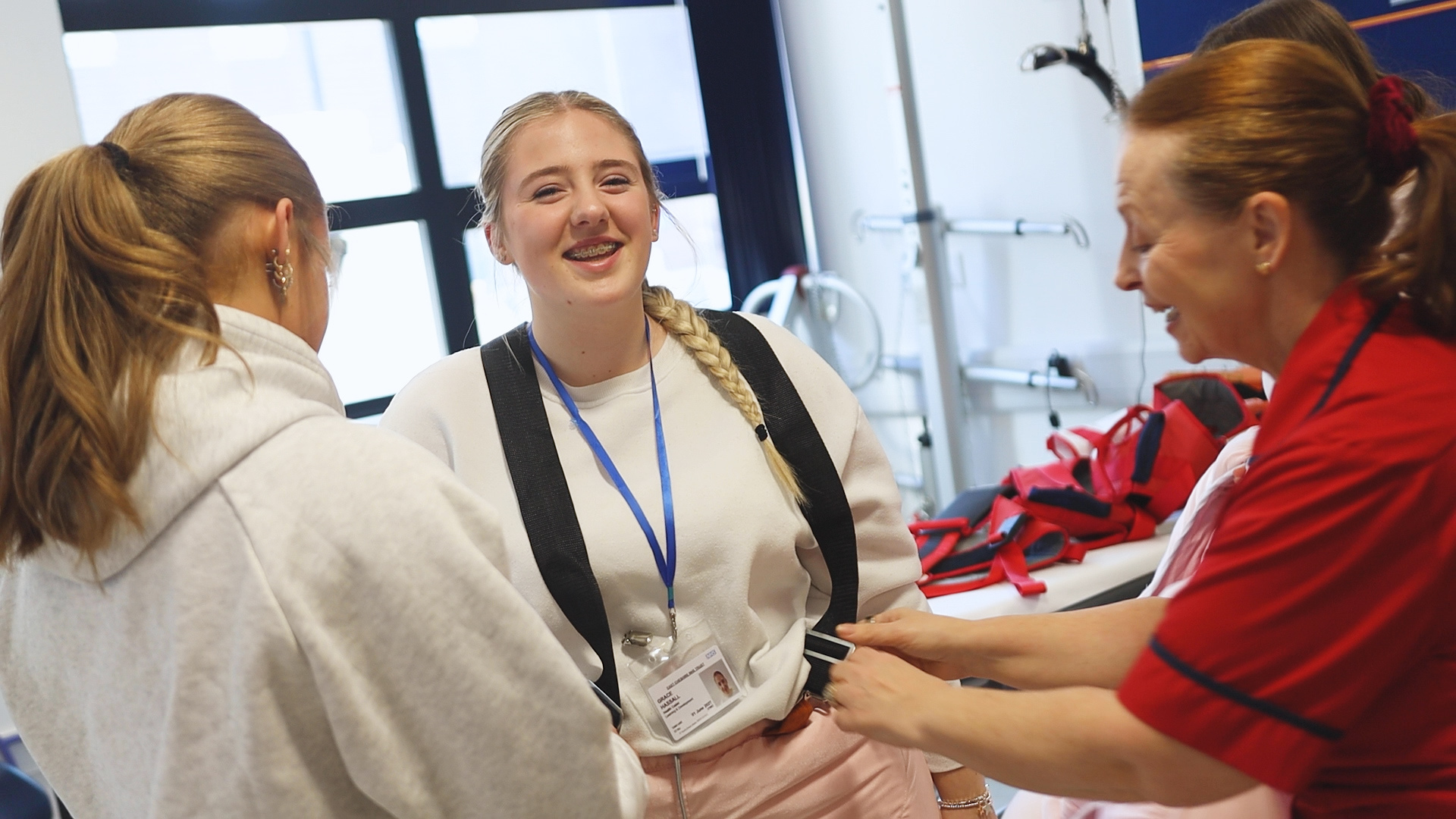 In a hospital setting, a hospital worker and a female health student are helping another health student into a fat suit.