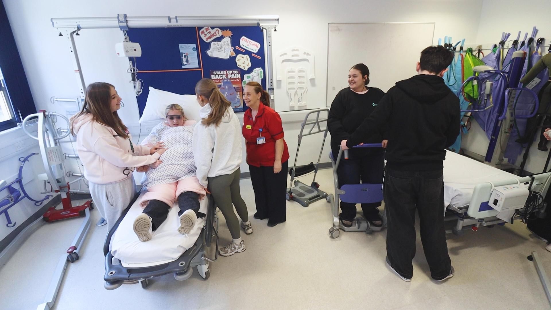 In a hospital setting, a young girl dressed in a fat suit lies on a bed while two other students and a hospital worker talk to her. Also in the room, two students are practising using a walking frame.