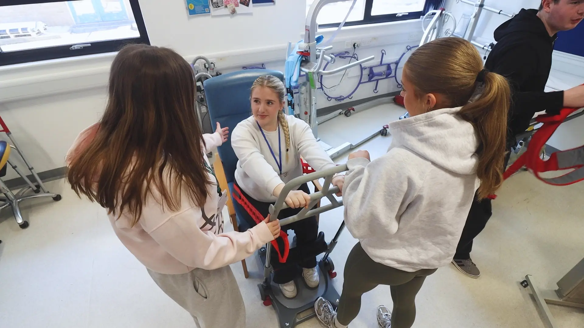 In a hospital setting, a young female sits on a chair with a walking frame attached to it. While two health students stand next to her.